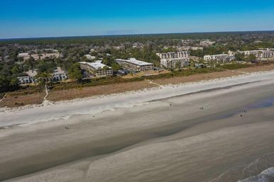Aerial view of a wide sandy beach with gentle waves, a dune path and rows of beachfront condos and trees under a clear blue sky