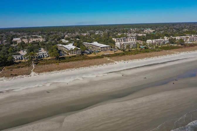 Aerial view of a wide sandy beach with gentle waves, a dune path and rows of beachfront condos and trees under a clear blue sky