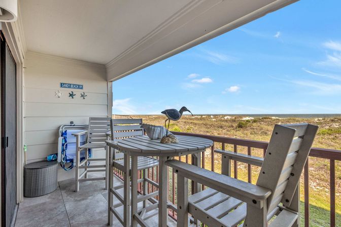 Beachfront balcony with high-top table and chairs, decorative shorebird centerpiece, coastal dunes and ocean view under a bright blue sky.