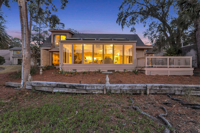Cozy dusk exterior of a suburban home featuring a glowing sunroom with floor-to-ceiling windows, an adjoining wooden deck, landscaped yard with pine mulch and a low timber retaining wall, framed by mature trees.