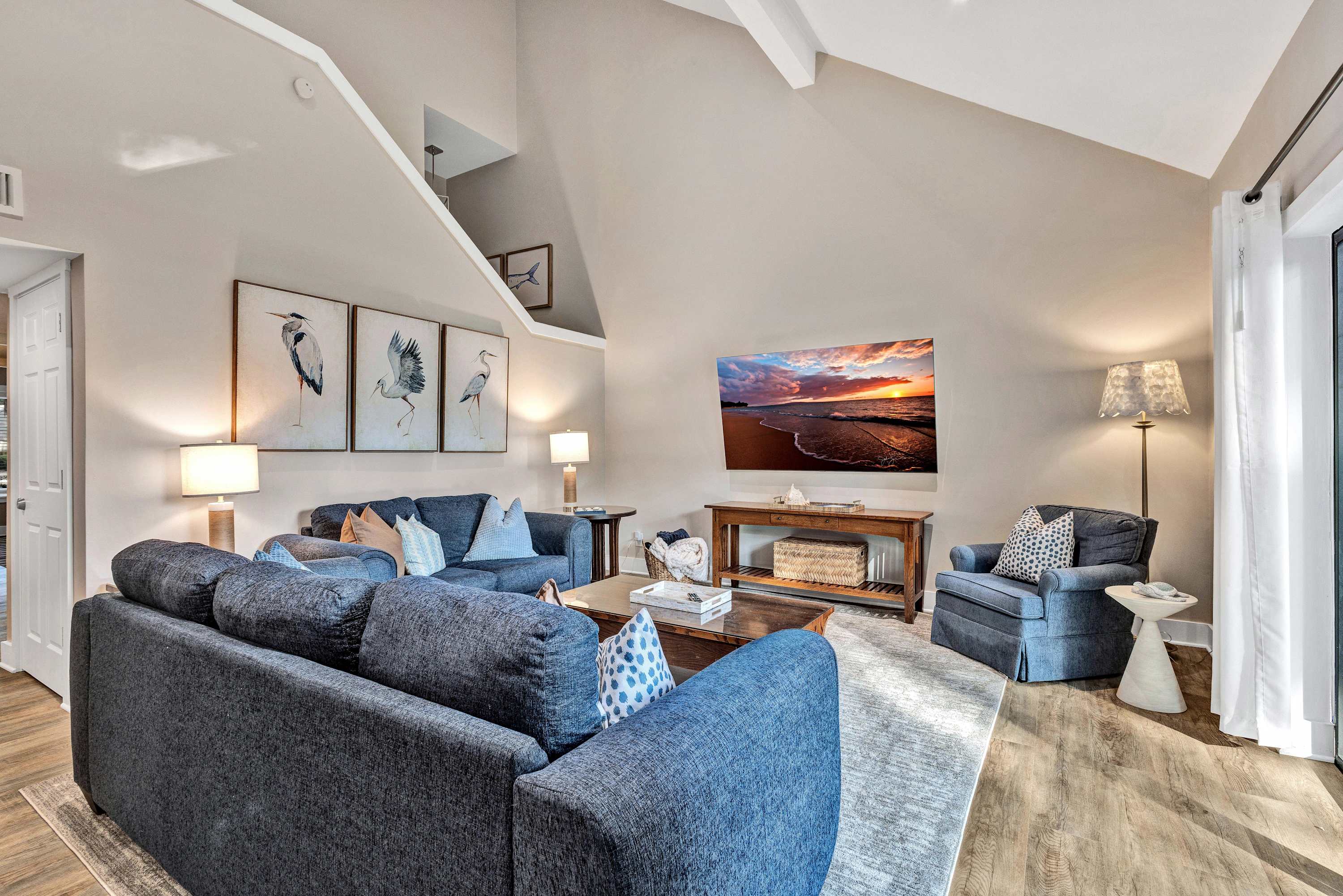 Cozy coastal-inspired living room with vaulted ceiling and natural light, navy blue sofas and armchair, hardwood floors, bird artwork, wooden coffee table and console, and a wall-mounted TV showing a beach sunset.
