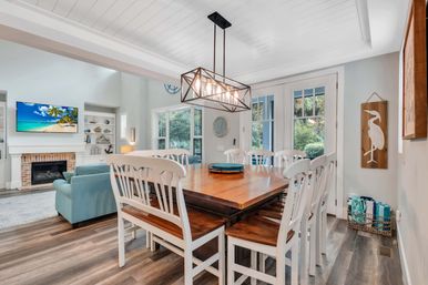 Bright coastal dining room in a beach-cottage style with a large wooden farmhouse table and white chairs under a geometric pendant light, French doors to greenery and an adjoining living area with blue sofa, fireplace and wall-mounted TV showing a tropical beach.
