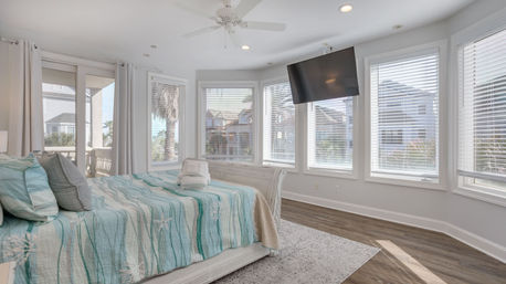 Sunlit coastal master bedroom with panoramic bay windows, aqua starfish quilted bed with folded towels, wall-mounted TV, ceiling fan and balcony view of palm trees and nearby beach houses.