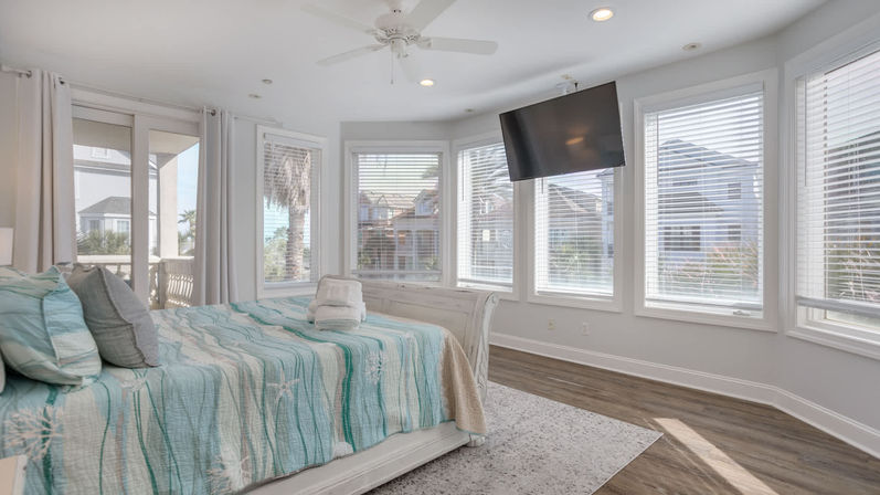 Sunlit coastal master bedroom with panoramic bay windows, aqua starfish quilted bed with folded towels, wall-mounted TV, ceiling fan and balcony view of palm trees and nearby beach houses.