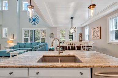Bright coastal open-concept kitchen and living area in beach-cottage style with a marble island and sink, pendant lights, turquoise sectional, and white dining chairs
