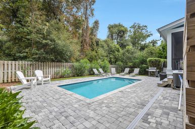 Rectangular in-ground backyard pool with blue water, gray paver patio, white Adirondack chairs and lounge chairs, gas grill near a screened porch, fenced yard backed by dense trees and greenery