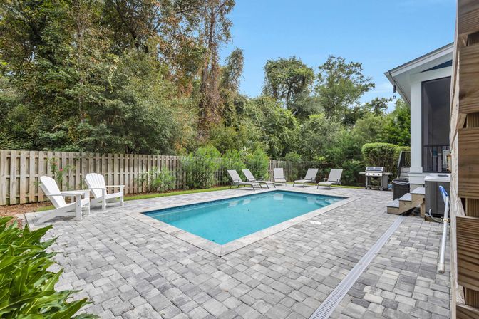 Rectangular in-ground backyard pool with blue water, gray paver patio, white Adirondack chairs and lounge chairs, gas grill near a screened porch, fenced yard backed by dense trees and greenery