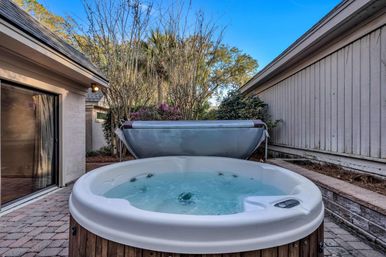 Bubbling outdoor hot tub on a suburban backyard patio with folded cover, paver stones, shrubs, and clear blue sky.