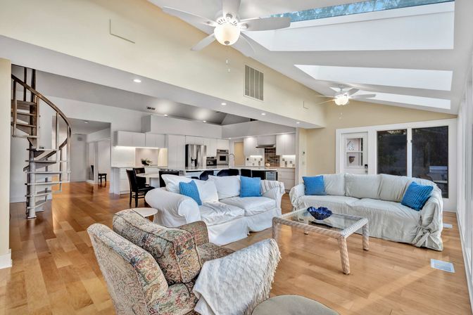 Bright, airy open-concept living room and kitchen with skylights and ceiling fans, hardwood floors, spiral staircase, white slipcovered sofas with blue pillows, glass coffee table and stainless-steel appliances