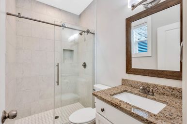 Bright modern residential bathroom with glass sliding shower, white marble-look tile, built-in shower bench, small hexagon tile floor, granite vanity with undermount sink and wood-framed mirror.