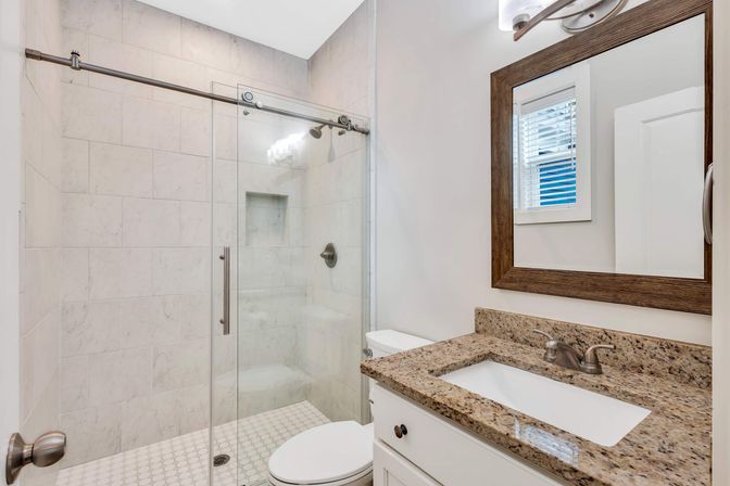 Bright modern residential bathroom with glass sliding shower, white marble-look tile, built-in shower bench, small hexagon tile floor, granite vanity with undermount sink and wood-framed mirror.