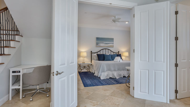 Open double doors reveal a coastal-style bedroom with a metal headboard, white bedding and navy pillows on a blue area rug, tile floors, matching bedside lamps and a small desk tucked under the stairs.