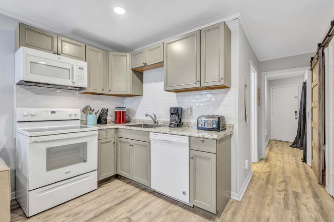 Bright compact kitchen with gray shaker cabinets, white stove, microwave and dishwasher, subway tile backsplash, granite countertops, light wood floors, coffee maker and toaster, and hallway entry