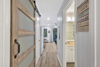 Bright coastal-style hallway in a beach-themed apartment with a sliding wood barn door, light wood floors, bathroom on the right and an open living area with balcony at the end.