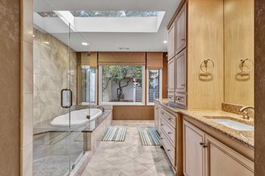 Spa-like luxury bathroom with skylight, glass-enclosed shower, built-in soaking tub, granite double vanity, marble tile floor, and large window framing a courtyard tree.
