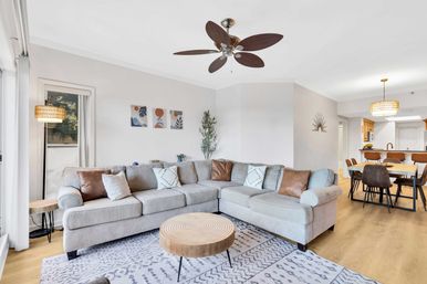 Bright, cozy modern open-plan living and dining area — light gray L-shaped sectional with leather accent pillows, round wooden coffee table on patterned rug, ceiling fan, floor lamp and adjacent wood dining table with pendant light.