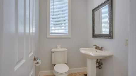 Compact bright powder room with white toilet and pedestal sink, brushed-nickel faucet, ornate framed mirror, neutral gray walls and a window with white blinds.