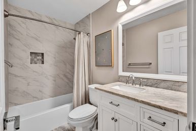 Contemporary residential bathroom with marble-look tile shower and recessed niche, curved shower rod with beige curtain, white tub and toilet, white vanity with marble countertop, large framed mirror and overhead sconces