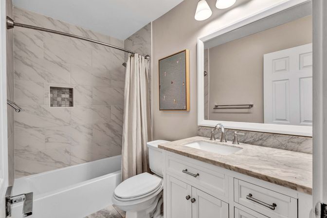Contemporary residential bathroom with marble-look tile shower and recessed niche, curved shower rod with beige curtain, white tub and toilet, white vanity with marble countertop, large framed mirror and overhead sconces