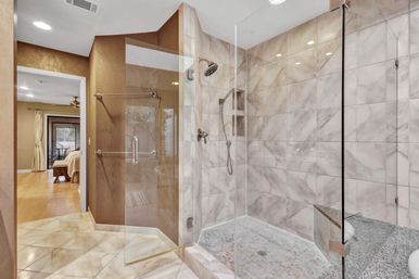 Luxury marble-tiled walk-in shower with glass enclosure, built-in bench, rainfall and handheld showerheads, open view into adjoining bedroom