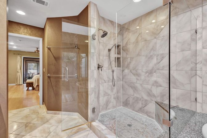 Luxury marble-tiled walk-in shower with glass enclosure, built-in bench, rainfall and handheld showerheads, open view into adjoining bedroom