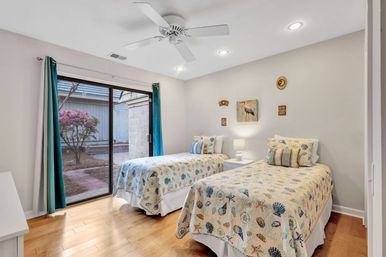 Coastal-themed guest bedroom with two twin beds in seashell quilts, white nightstand and lamp, hardwood floors, ceiling fan and teal curtains framing a sliding glass door to a small courtyard with a flowering shrub.