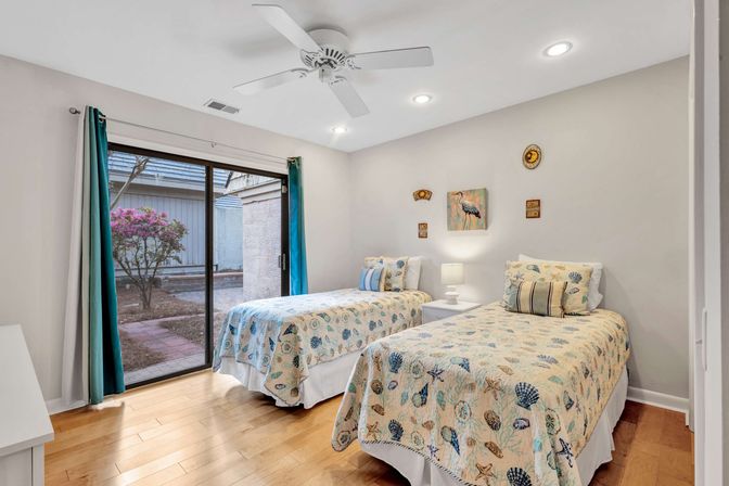 Coastal-themed guest bedroom with two twin beds in seashell quilts, white nightstand and lamp, hardwood floors, ceiling fan and teal curtains framing a sliding glass door to a small courtyard with a flowering shrub.