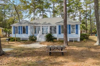 Blue-shuttered coastal cottage with white steps and glass sunroom entrance, nestled among tall pine trees on a sandy yard with a picnic table and small charcoal grill.