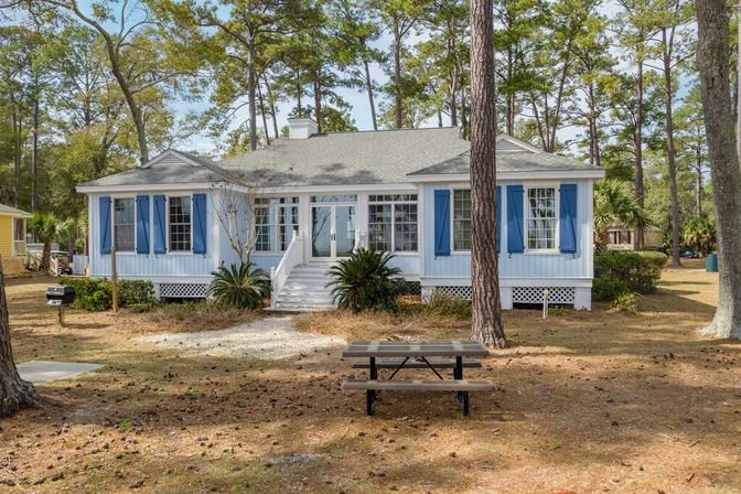 Blue-shuttered coastal cottage with white steps and glass sunroom entrance, nestled among tall pine trees on a sandy yard with a picnic table and small charcoal grill.