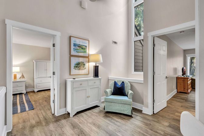 Bright modern entryway with high ceiling and wood-look floors, mint-green accent chair with navy pillow in a cozy sitting nook beside a white console topped with a lamp and framed coastal prints, open doorways leading to bedrooms with white furniture.