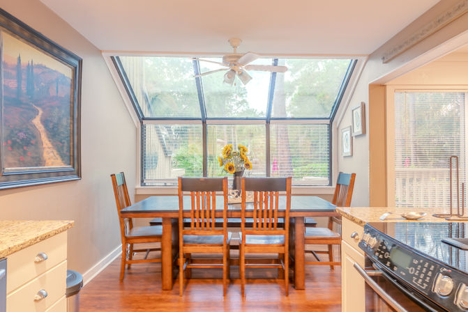 Bright dining room with wooden table and six chairs under slanted skylight windows, sunflower centerpiece, ceiling fan, hardwood floors and nearby kitchen island with stove.
