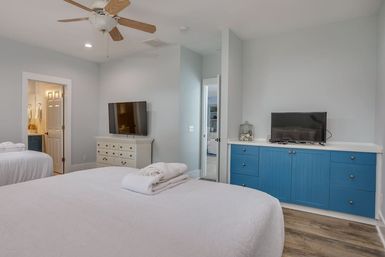 Bright coastal-style bedroom with crisp white bedding on two beds, wood ceiling fan, wall-mounted TVs, blue built-in cabinets, hardwood floors and visible en-suite bathroom.