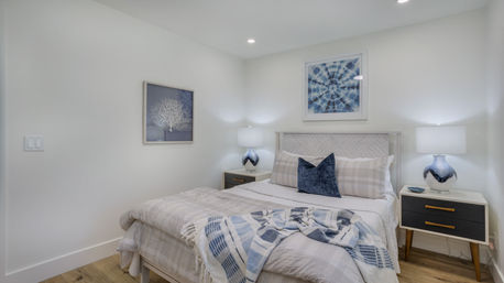 Bright modern bedroom with neutral plaid bedding, blue throw and velvet pillow, matching blue ceramic lamps on mid-century nightstands, coastal-inspired blue artwork above a white wood headboard, white walls and light hardwood floor.