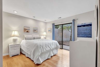 Bright coastal-style bedroom with white linens on a bed, hardwood floors, two bedside lamps, blue curtains, sliding glass patio door and ocean-themed wall art.