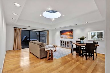 Sunlit open-concept living and dining room with round skylight, honey hardwood floors, beige sofa, wall-mounted TV, sliding glass doors and wooden dining table.