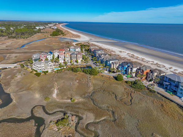 Aerial shot of pastel beachfront homes along a curving sandy shoreline, surrounded by tidal marshes and blue ocean under a clear sunny sky.