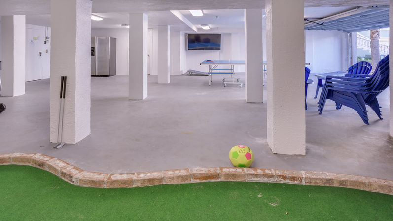 Basement indoor recreation room with concrete floor and artificial putting green in the foreground, bright neon soccer ball, golf putters leaning by a column, ping-pong table, stacked blue plastic chairs and a stainless refrigerator in the back.