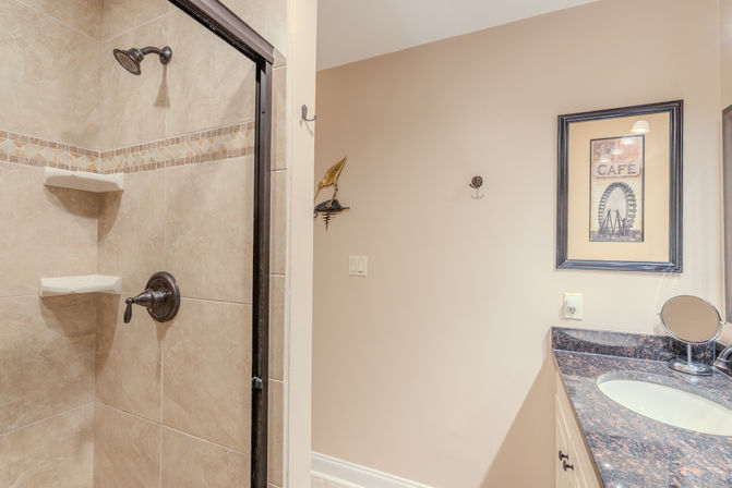 Beige-tiled shower with oil-rubbed bronze fixtures and corner shelves beside a granite-topped vanity and sink, mirror and framed cafe print in a residential bathroom interior.