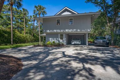 Raised modern coastal home with carport and two parked cars, shaded concrete driveway framed by palm trees and lush greenery on a sunny day.