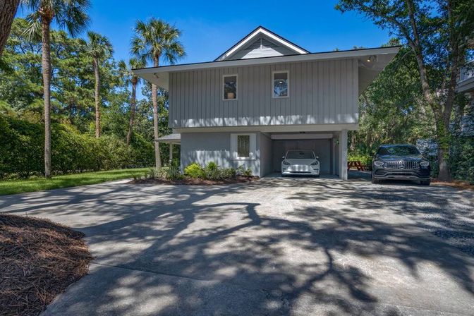 Raised modern coastal home with carport and two parked cars, shaded concrete driveway framed by palm trees and lush greenery on a sunny day.