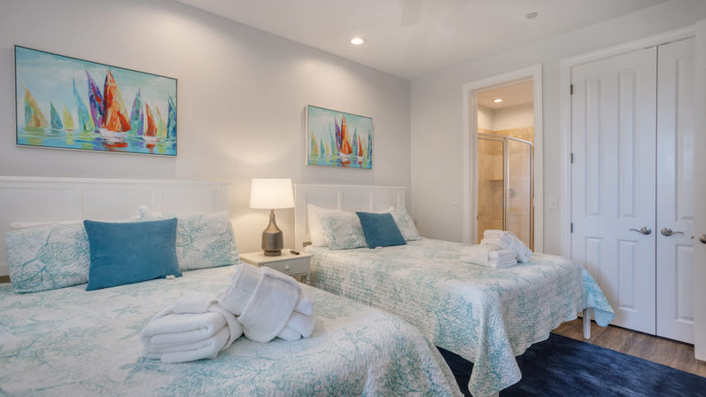 Coastal-themed guest bedroom with two beds in teal coral quilts, blue accent pillows, a bedside lamp, folded white towels, sailboat paintings, and an en-suite shower.