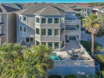 Three-story coastal vacation condo with wraparound windows, stacked balconies, palm trees and a sunny fenced patio with lounge chairs and a small plunge pool.