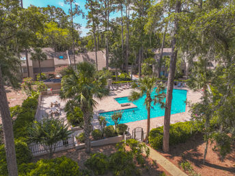 Aerial view of a community swimming pool with bright turquoise water, pool deck with lounge chairs and picnic tables, fenced gate, palm trees and tall pines, and townhouses tucked into a wooded neighborhood.
