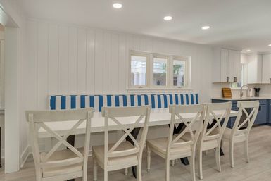 Bright coastal-style kitchen dining nook with white shiplap walls, blue-and-white striped banquette, long white table and six cross-back chairs, open to modern navy and white kitchen