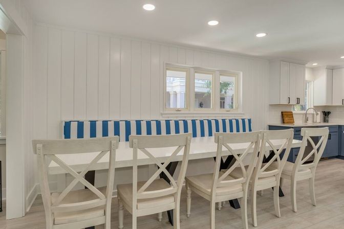 Bright coastal-style kitchen dining nook with white shiplap walls, blue-and-white striped banquette, long white table and six cross-back chairs, open to modern navy and white kitchen