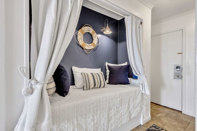 Cozy nautical-inspired built-in daybed nook with navy accent wall, white tie-back curtains, striped and navy throw pillows, overhead brass wall sconce, and decorative rope lifebuoy