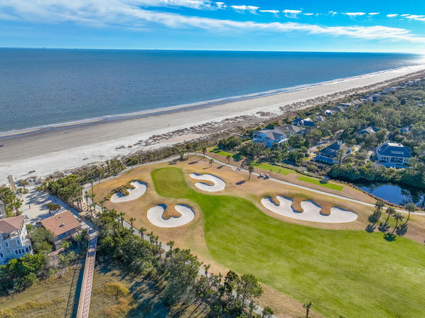Aerial view of an oceanfront golf course with bright green fairways and curvy white sand bunkers beside a wide sandy beachfront and seaside homes under a bright blue sky.