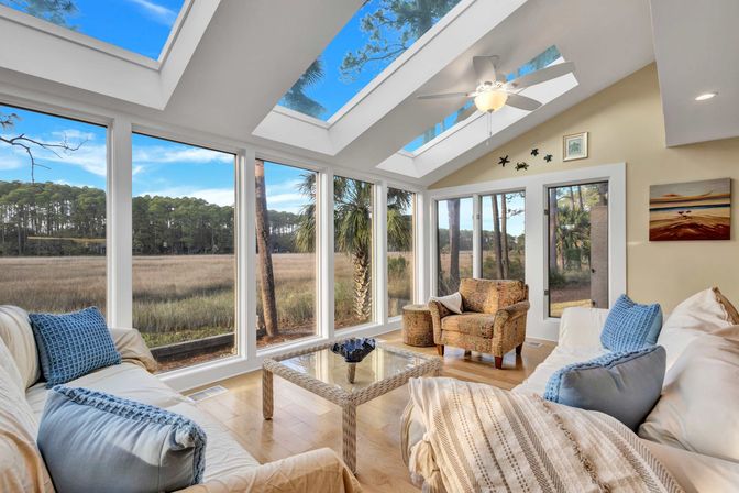 Sun-drenched coastal sunroom living room with skylights and floor-to-ceiling windows overlooking tidal marsh and pine trees, neutral sofas with blue pillows, patterned armchair and glass coffee table.
