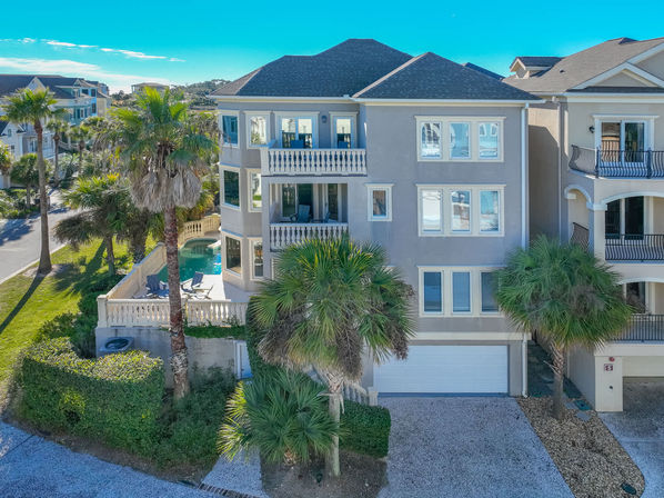 Sunlit three-story coastal vacation home with balconies, private pool, palm trees and garage set among neighboring beach houses under a clear blue sky