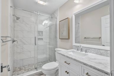 Bright spa-like modern bathroom with frameless glass walk-in shower, marble-effect tiled walls and mosaic shower floor, white vanity with marble countertop, chrome fixtures and large mirror.